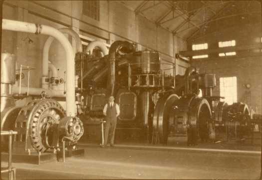 Shows the generating equipment inside the Power House. Unidentified man posing for the photograph.