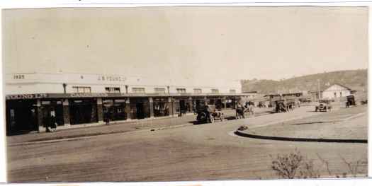Shows shops to the left and a variety of vehicles on the street. Red Hill in background.