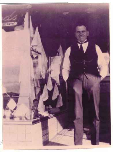 Taylor is standing at the entrance of his shop. The word 'Costumier' is on the window to the left.