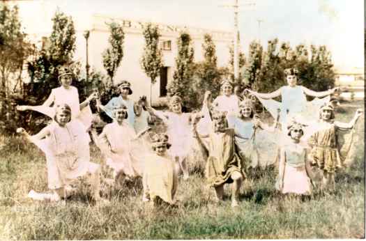 JB Young original store can be seen behind the trees. Hand tinted photograph of about 12 young girls dressed up.