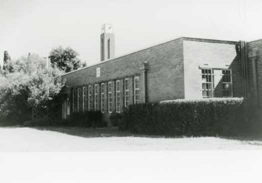 Single storey brick office with clock tower, trees and hedge. See notes.